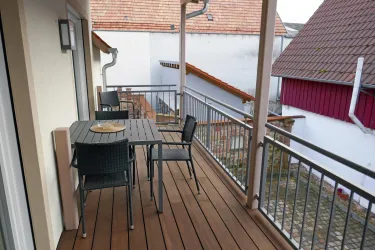 Ein Balkon mit einem Holztisch und mehreren Stühlen. Im Hintergrund sind angrenzende Dächer und Wände zu sehen.