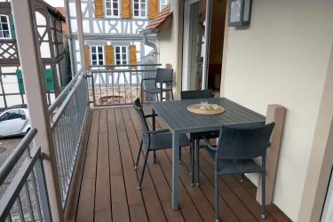 Ein gemütlicher Balkon mit einem Tisch und Stühlen aus Metall. Im Hintergrund sind traditionelle Fachwerkhäuser zu sehen.