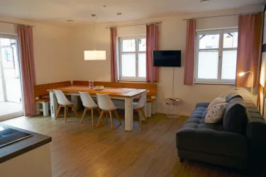 Ein helles Wohnzimmer mit einem modernen Esstisch und Stühlen. Rechts steht ein bequemes Sofa, und große Fenster lassen viel Licht herein.