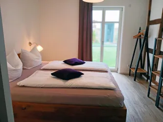 Ein gemütliches Schlafzimmer mit einem Doppelbett und lila Kissen. Ein großer Fensterbereich sorgt für viel Tageslicht und Blick ins Grüne.