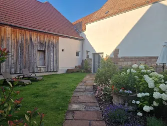 Ein schöner Garten mit einem Steinweg und bunten Blumen. Im Hintergrund ist ein Haus mit Holzfassade und ein klarer blauer Himmel zu sehen.