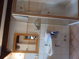 Ein modernes Badezimmer mit einer Dusche, einem Waschbecken und einem Spiegel. Die Wände sind hell gefliest und der Boden ist aus keramischen Fliesen.
