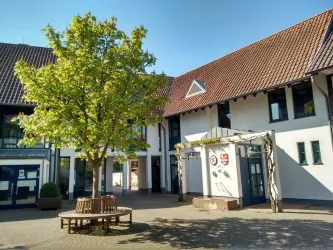 Ein ruhiger Innenhof mit einem großen Baum und einer Sitzbank aus Holz. Die Gebäude im Hintergrund haben weiße Wände und rote Dächer.