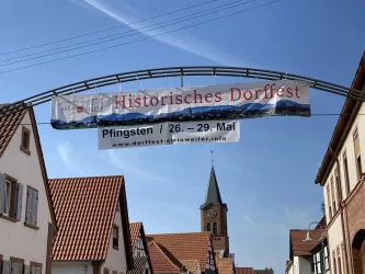 Ein historisches Dorffest mit einem großen Banner über der Straße. Im Hintergrund ist eine Kirche und hübsche, traditionelle Gebäude zu sehen.