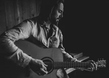 Ein Mann sitzt und spielt Gitarre in einem dunklen Raum. Der Fokus liegt auf seiner konzentrierten Miene und der Instrument.