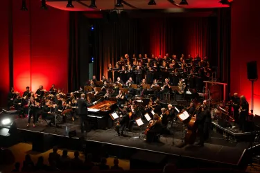 Ein Orchester und ein Chor treten auf einer Bühne mit roter Beleuchtung auf. Die Musiker konzentrieren sich auf ihre Instrumente und die Performance.