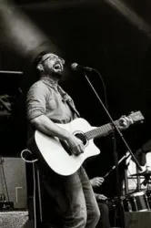 Ein Musiker singt leidenschaftlich in ein Mikrofon und spielt Gitarre. Die Bühne ist gut beleuchtet und die Atmosphäre ist lebhaft.