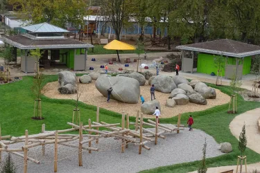 Ein Spielplatz mit großen Steinen und Holzgerüsten, umgeben von Rasen und Sand. Im Hintergrund sind Gebäude und ein gelbes Sonnensegel sichtbar.