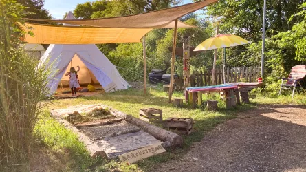 Ein gemütlicher Campingplatz mit einem Tipi-Zelt und einer Sitzgelegenheit aus Holz. Sonnensegel spenden Schatten in einer ruhigen, grünen Umgebung.