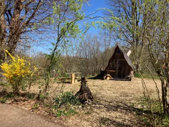 Ein kleines, gemütliches Häuschen in der Natur, umgeben von frischen Bäumen und Pflanzen. Der Himmel ist blau und die Landschaft ist ruhig und einladend.