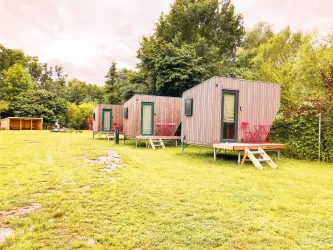 Drei moderne, graue Ferienhäuser stehen auf einer grünen Wiese. Umgeben von Bäumen und mit pinken Stühlen auf der Veranda bieten sie einen gemütlichen Rückzugsort.