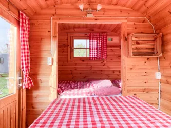Ein gemütlicher Raum aus Holz mit einem großen Bett und gestreiftem Tischdeckenmuster. Die Fenster sind mit roten Vorhängen geschmückt und bieten einen Blick nach draußen.