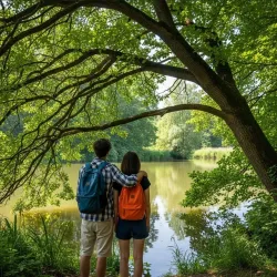Ein Paar steht am Ufer eines ruhigen Sees, umgeben von dichten, grünen Bäumen. Die beiden haben Rucksäcke dabei und schauen entspannt auf das Wasser.