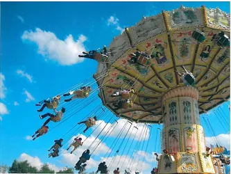 Ein bunter Freizeitpark mit einem Schwungkarussell, auf dem viele Menschen fröhlich schwingen. Der Himmel ist klar und blau, mit wenigen Wolken.