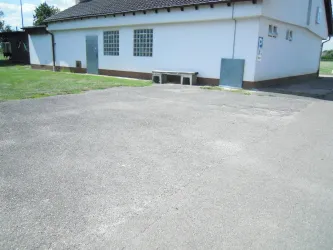 Ein leerer, asphalierter Platz neben einem weiß gestrichenen Gebäude. Im Hintergrund sind grüne Wiesen und ein blauer Himmel zu sehen.