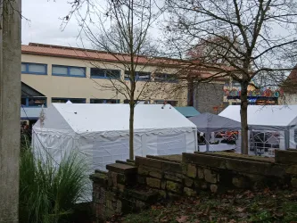 Ein kleiner Markt mit mehreren weißen Zelten ist zu sehen. Im Hintergrund ist ein Gebäude und einige Bäume sichtbar.