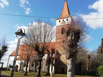 Eine alte Kirche mit einem hohen, spitzen Turm steht vor einem blauen Himmel. Um die Kirche herum sind kahl geschnittene Bäume und eine Straßenlaterne zu sehen.