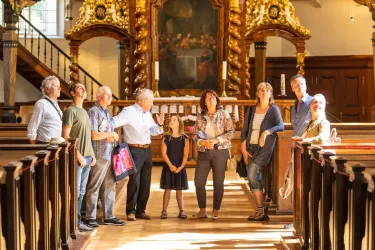 Eine Gruppe von Menschen steht in einer Kirche und schaut neugierig nach oben. Die goldene Ausstattung und die Holzbankreihen schaffen eine warme Atmosphäre.