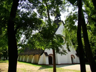 Eine weiße Kirche umgeben von hohen Bäumen. Der Himmel ist klar und die Umgebung wirkt friedlich.