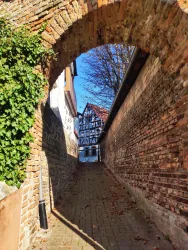 Ein schmaler Weg, der durch einen Bogen aus Ziegelsteinen führt. Im Hintergrund ist ein Fachwerkhaus sichtbar, umgeben von blauen Himmel und Bäumen.