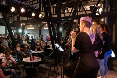 Eine Sängerin steht auf der Bühne und singt in einem lebhaften Veranstaltungsraum. Das Publikum sitzt an Tischen und ist aufmerksam.