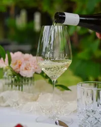 Ein elegantes Weinglas wird mit Weißwein gefüllt. Im Hintergrund sind zarte Blumen und Dekorationen zu sehen.