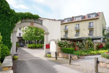 Ein einladender Eingang zu einem Hotel mit einem blühenden Garten. Das Gebäude ist umgeben von Bäumen und bietet eine entspannte Atmosphäre.