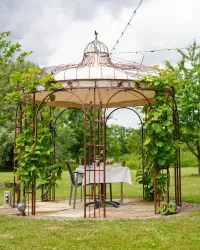 Ein hübsches Gartenhaus mit einem Tisch und Stühlen, umgeben von Pflanzen. Die Szenerie ist grün und einladend, ideal für entspannte Momente im Freien.