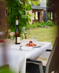 Ein elegant gedeckter Tisch im Freien mit einem Glas Wein und einer kleinen Dessert-Anrichtung. Umgeben von einer grünen Gartenlandschaft sorgt die Szene für eine einladende Atmosphäre.
