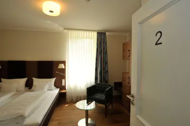 Ein modernes Hotelzimmer mit einem Doppelbett und einem gemütlichen Sessel. Das Zimmer hat große Fenster mit Vorhängen und eine warmes Licht.