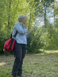 Eine Person steht im Freien und spricht, umgeben von Bäumen. Sie trägt eine graue Bluse und eine rote Tasche.