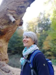 Eine Frau mit grauen Haaren steht in einem Wald und lächelt. Im Hintergrund sind Bäume mit herbstlichen Farben zu sehen.
