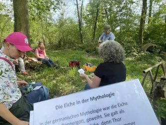 Eine Gruppe von Menschen sitzt im Wald und diskutiert unter einem Baum. Im Vordergrund ist ein Informationsblatt über die Eiche in der Mythologie sichtbar.