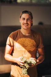 Ein junger Mann mit einem lächelnden Gesicht steht drinnen und hält Weintrauben in der Hand. Er trägt ein braunes T-Shirt und hat eine lässige Ausstrahlung.