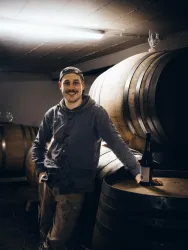 Ein Weinbauer steht lächelnd neben großen Holzfässern in einem Weinkeller. Er hält eine Flasche Wein in der Hand.