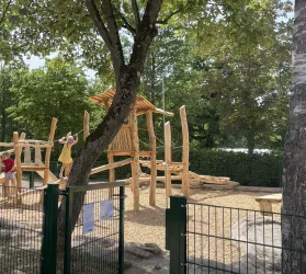 Ein Spielplatz aus Holz mit verschiedenen Kletterelementen und Rutsche. Die Umgebung ist grün und von Bäumen umgeben.
