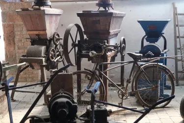 Eine Sammlung alter Mühlentechnik steht in einem Raum. Im Vordergrund ist ein Fahrrad und mehrere Mühlen mit großen Trichtern zu sehen.
