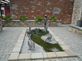 Eine Bronzeskulptur eines Jungen mit einem Brunnen und mehreren Enten um ihn herum. Der Platz ist mit Steinen und blühenden Pflanzen gestaltet.
