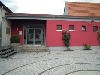 Ein modernes Gebäude mit roter Fassade und großen Fenstern. Vor dem Eingang befindet sich ein gepflasterter Vorplatz mit grünen Pflanzen.