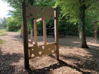 Ein Spielgerät im Freien aus Holz mit durchbrochenen Formen. Der Bereich ist von Bäumen umgeben und auf dem Boden liegen Holzspäne.