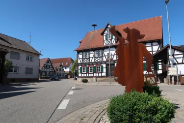 Eine malerische Straße mit traditionellen Fachwerkhäusern und klarem blauen Himmel. Im Vordergrund steht eine kunstvolle Figur.