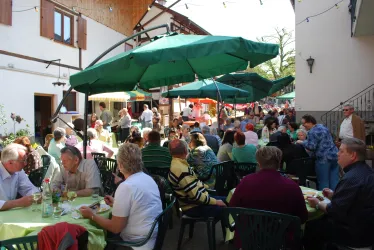 Eine lebhafte Veranstaltung im Freien mit vielen Gästen, die an Tischen sitzen. Der Raum ist mit grünen Sonnenschirmen und bunten Dekorationen geschmückt.