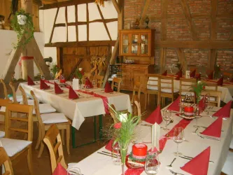 Ein gemütlicher Speisesaal mit mehreren Tischen, die mit weißen Tischdecken und roten Servietten gedeckt sind. Im Hintergrund befinden sich rustikale Holzelemente und eine Holzvitrine.