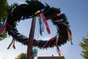 Ein großer Kranz mit bunten Bändern hängt an einem Holzpfahl. Der Himmel ist klar und blau.