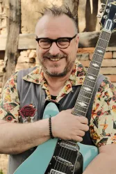 Ein lächelnder Mann mit Brille hält eine blaue Gitarre. Er trägt ein farbenfrohes Hemd und steht vor einer rustikalen Hintergrundkulisse.