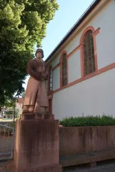 Eine Statue einer Person in traditioneller Kleidung steht vor einem Gebäude. Die Statue ist aus Stein und von Bäumen umgeben.