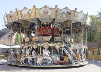 Ein nostalgisches Karussell mit bunten Figuren und dekorativen Elementen steht auf einem Platz. Es ist ein sonniger Tag und die Umgebung wirkt einladend.
