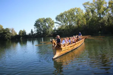 Eine Gruppe von Menschen fährt in einem Holzboot auf einem ruhigen Gewässer. Umgeben von grünen Bäumen und klarem Himmel genießen sie die Natur.