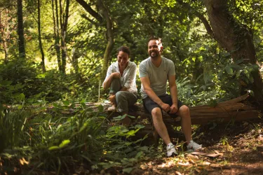 Ein Paar sitzt entspannt auf einem Baumstamm im Wald. Die grüne Umgebung wirkt ruhig und einladend.