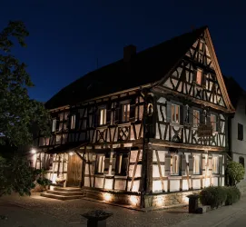 Ein historisches Fachwerkhaus bei Nacht, beleuchtet und einladend. Es hat grüne Pflanzen und einen gepflasterten Weg vor der Tür.